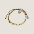 Pulsera Cabrera con cuentas de crisoprasa verde limón, separadores dorados, cordón de macramé marrón ajustable y dije de lazo solidario en acero inoxidable dorado, sobre fondo neutro.